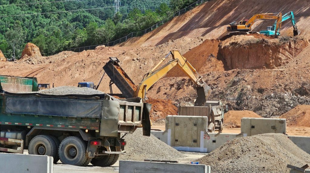 Tiến độ xây dựng Dự án Vinhomes Làng Vân Tháng 8/2025: Bứt phá mạnh mẽ với quy mô khủng 4 Cảnh thi công tại khu đầu đường dẫn vào suối Lương, dự án Vinhomes Làng Vân, với máy xúc đang bốc đất đá lên xe tải, phía sau là sườn đồi đã được đào xẻ, bao quanh bởi rừng cây xanh.